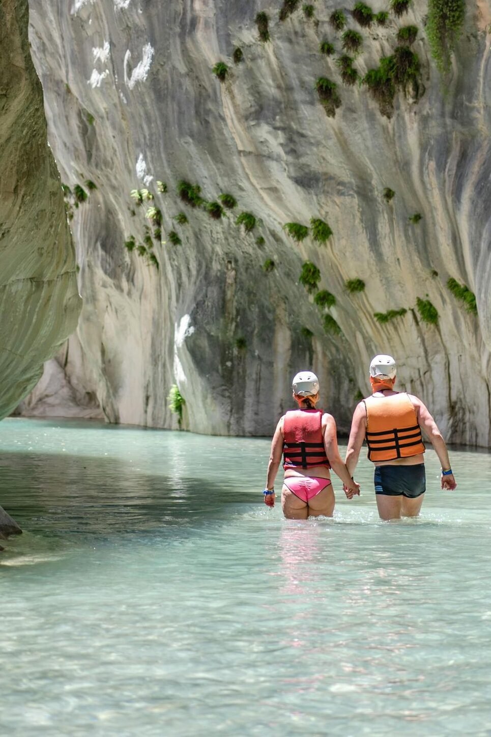 Two adults explore a scenic canyon, wearing life jackets in clear waters, surrounded by rocky cliffs.