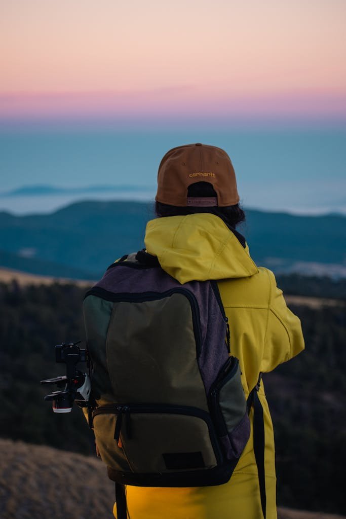 Female hiker in yellow jacket with backpack enjoying a scenic sunset view from a hilltop.